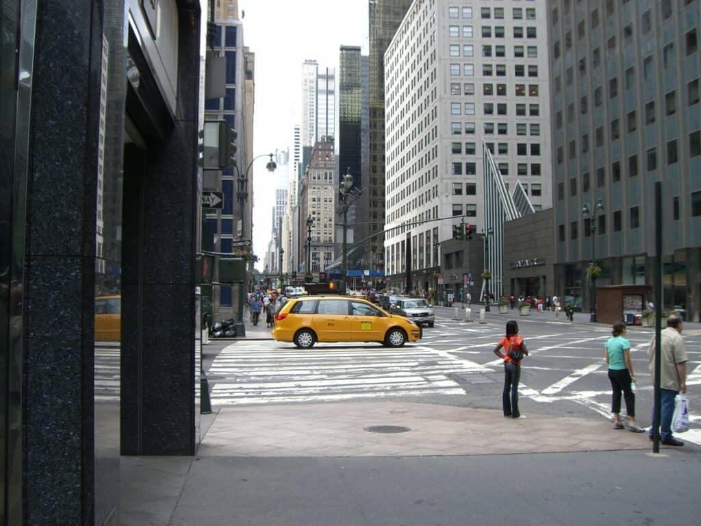 En gatukorsning i New York, människor står och väntar vid ett övergångsställe, en gul taxibil står på ett annat övergångsställe. Höga byggnader åt alla håll och himlen skymtar.