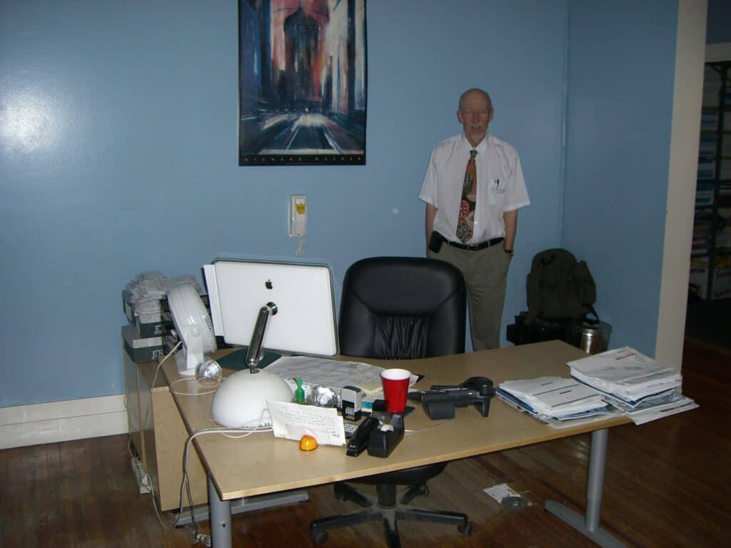 Kontor med skrivbord, dator, skrivbordsstol. Bakom står Rehabilitation Internationals generalsekreterare Tomas Lagerwall i vit skjorta, slips och beigea byxor. Väggen bakom är ljusblå, med en tavla och en telefon mitt på väggen.